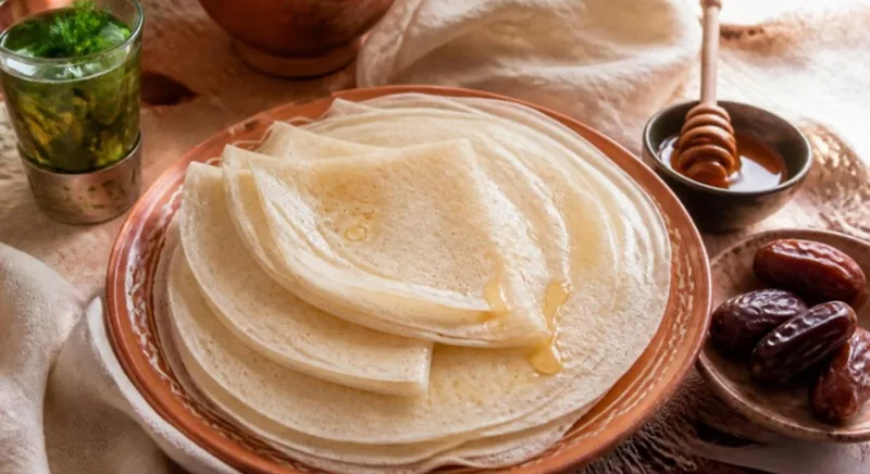 Crêpes sans gluten à la farine de riz, fines et claires, pliées sur un plat en terre cuite avec du miel, du thé à la menthe et des dattes
